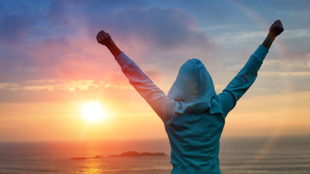 A hooded figure holds their arms up in victory in front of an oceanside sunside.