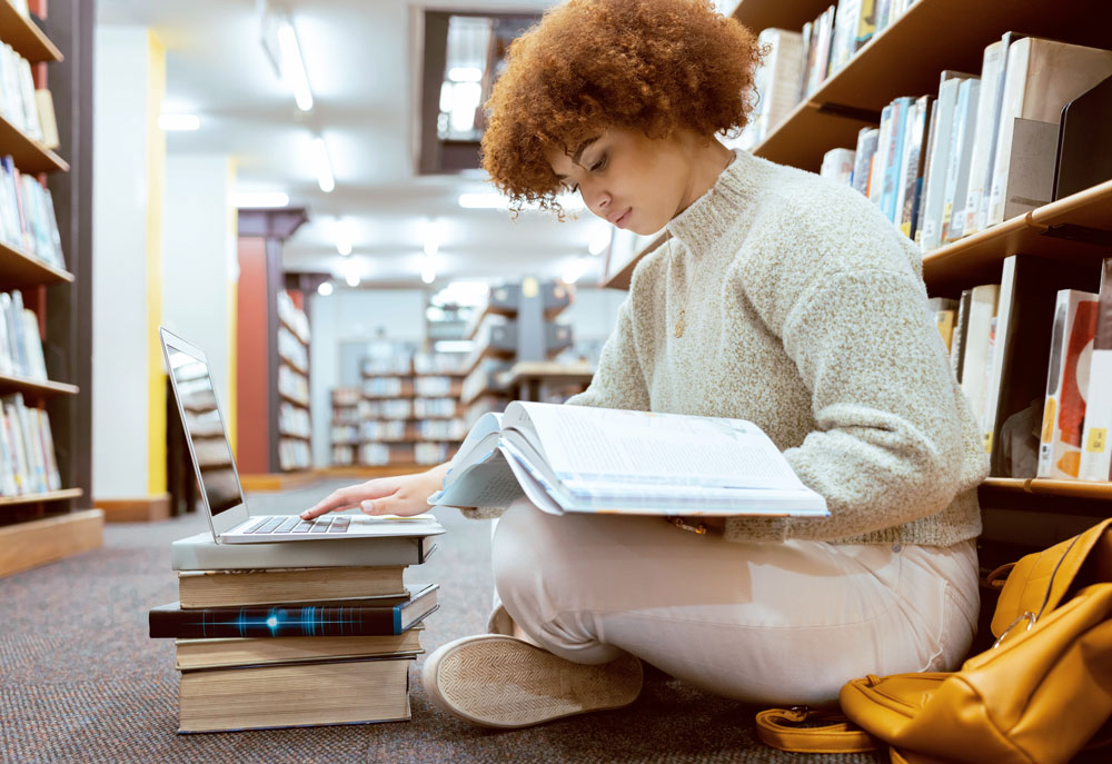 A prospective RBT reads from a book and her laptop while studying in a library.